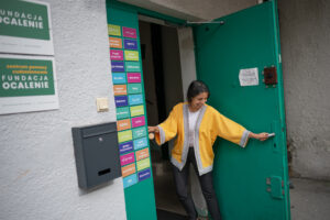 Mentorka kulturowa z uśmmiechem otwiera drzwi wejśćiowe Centrum Pomocy Cudzoziemcom w Warszawie. Na drzwiach napis "cześć" w kilkunastu językach. Na ścianie budynku tabliczki z logotypami Fundacji.