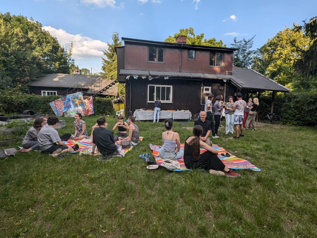Piknik w parku na Jazdowie, Grupa ludzi rozmawia - część siedzi na kocach na trawie, część stoi w mniejszych grupkach. Za nimi drewniany domek fiński Osiedla Jazdów.