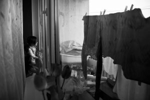 Czarno-biała fotografia. Dwie małe dziewczynki wyglądają przez okno. Widać rozwieszone na balkonie pranie.
