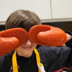 Chłopiec pozuje z silikonowymi rękawicami kuchennymi, zakrywając nimi oczy. Uśmiecha się. W tle przestrzeń kuchni.
